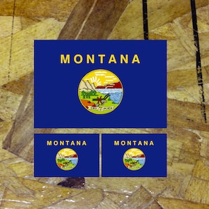 May include: Three blue and white Montana state flag stickers with the state name and seal. The seal features a mountain range, a sun, a river, and a plow.