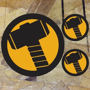 May include: Three black and yellow circular stickers with a silhouette of a hammer inside. The hammer is a symbol of Thor, the Norse god of thunder.