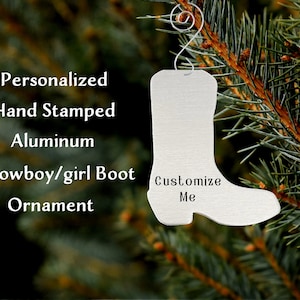 May include: A silver aluminum cowboy boot ornament with the text "Customize Me" on the side. The ornament is hanging from a pine tree branch.