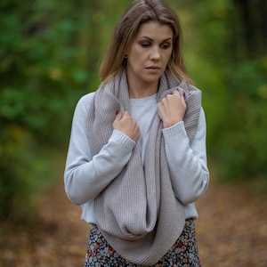 以下が含まれることがあります： 白いセーターと花柄のスカートを着た女性が、薄いグレーのニットのストールを巻いています。ストールは首に巻かれ、緩く結ばれています。