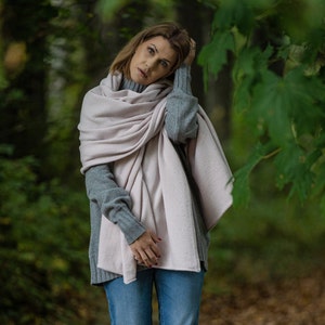 May include: A woman wearing a light pink scarf draped over a gray sweater and blue jeans. The scarf is wrapped around the neck and shoulders. The background is a blurred view of a forest.