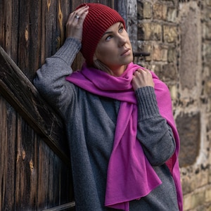 Könnte beinhalten: Eine Frau trägt einen grauen Pullover und einen rosa Schal mit einer roten Strickmütze. Die Frau steht vor einer Holzwand.