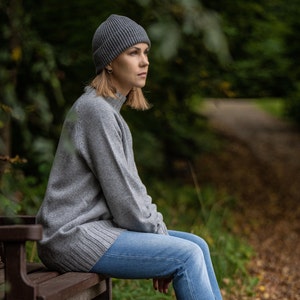 May include: A woman wearing a gray beanie and a gray sweater sits on a wooden bench. She is wearing blue jeans and has her legs crossed. The background is a blurred out forest.