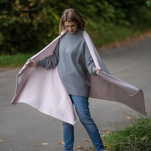 May include: A woman wearing a gray sweater and blue jeans is holding a reversible blanket. The blanket is light pink on one side and a lighter shade of pink on the other. The woman is standing on a paved path with green foliage in the background.