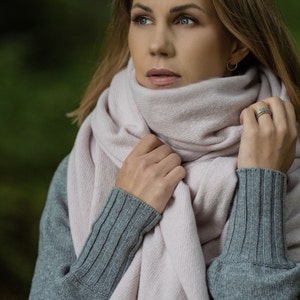 May include: A light pink scarf wrapped around the neck, paired with a gray sweater. The scarf is soft and drapes loosely. The sweater has ribbed cuffs. The background is a blurred green.