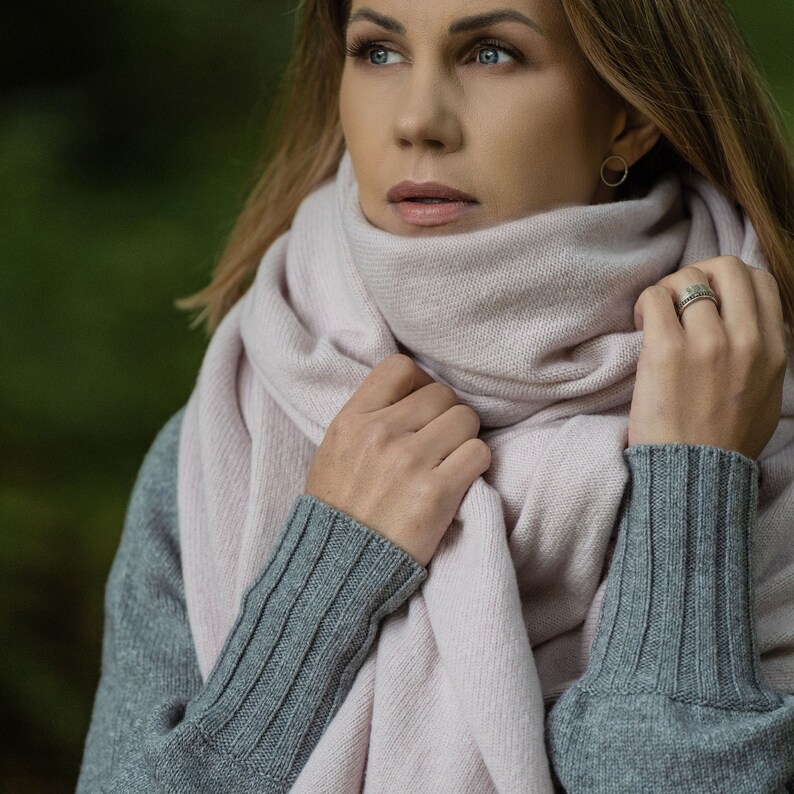 May include: A light pink scarf wrapped around the neck, paired with a gray sweater. The scarf is soft and drapes loosely. The sweater has ribbed cuffs. The background is a blurred green.