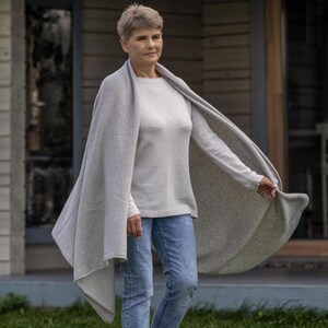 May include: A woman wearing a light gray sweater, blue jeans, and a matching light gray shawl. The shawl is draped over the shoulders and arms, with one side flowing in the wind. The background shows a house with a wooden exterior.