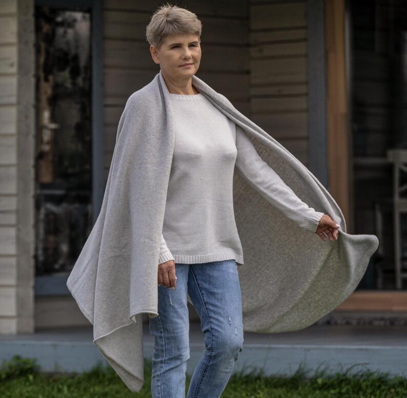 May include: A woman wearing a light gray sweater, blue jeans, and a matching light gray shawl. The shawl is draped over the shoulders and arms, with one side flowing in the wind. The background shows a house with a wooden exterior.