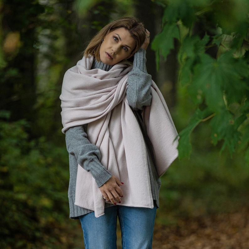 May include: A woman wearing a light pink scarf draped over a gray sweater and blue jeans. The scarf is wrapped around the neck and shoulders. The background is a blurred view of a forest.