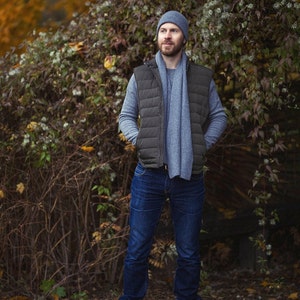 May include: A man wearing a gray beanie, a gray scarf, a black puffer vest, and blue jeans. He is standing in front of a leafy green bush.