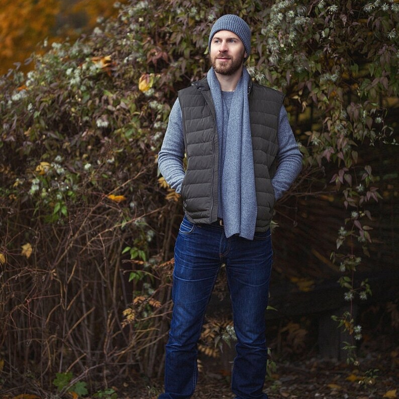 May include: A man wearing a gray beanie, a gray scarf, a black puffer vest, and blue jeans. He is standing in front of a leafy green bush.