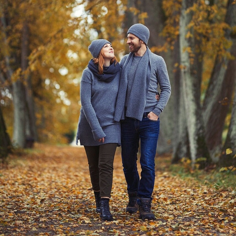 May include: A couple wearing gray knit hats and scarves walk through a forest. The woman is wearing a gray sweater and the man is wearing a gray sweater and jeans.