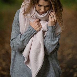 May include: A person wearing a light gray sweater and a pale pink scarf. The scarf is wrapped around the neck and held in place by the hands. The sleeves of the sweater have ribbed cuffs.