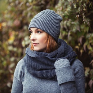 May include: A woman wearing a gray knitted hat and a matching gray scarf. The hat is a beanie style and the scarf is a long, thin scarf that is wrapped around her neck.