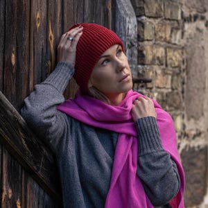 Puede incluir: Una mujer con un gorro de punto rojo y una bufanda rosa. Está de pie frente a una puerta de madera y una pared de piedra.