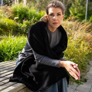 May include: A black shawl draped over a gray sweater. The shawl is wrapped around the shoulders and chest. The person is sitting on a wooden bench outdoors, with greenery in the background.