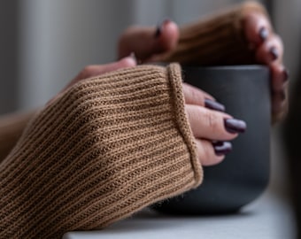 May include: A close-up of a person's hands wearing brown knitted fingerless gloves, holding a black mug.