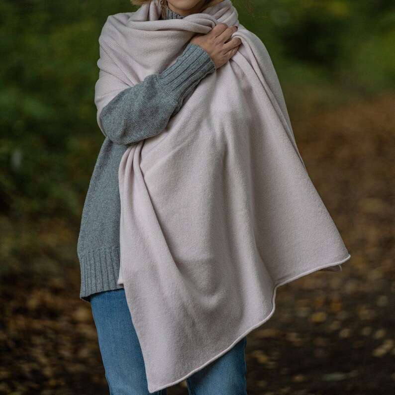 May include: A light pink shawl draped over a person wearing a gray sweater and blue jeans. The shawl is made of a soft, textured material. The background is blurred, with hints of green and brown.