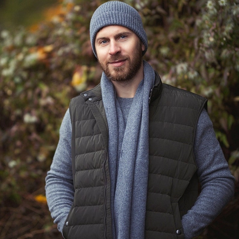 May include: A man wearing a gray knitted beanie, a gray scarf, and a dark green quilted vest. He is standing in front of a blurred background of green foliage.