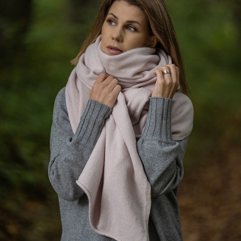May include: A woman wearing a light pink scarf and a gray sweater. The scarf is wrapped around the neck and shoulders. The background is a blurred green forest.