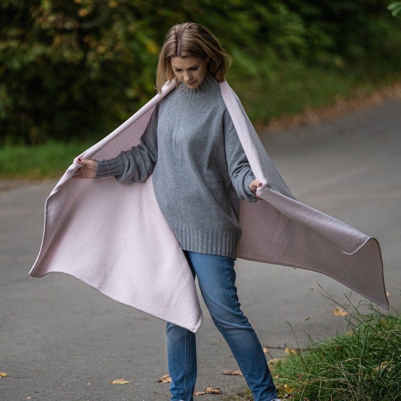 May include: A woman wearing a gray sweater and blue jeans is holding a reversible blanket. The blanket is light pink on one side and a lighter shade of pink on the other. The woman is standing on a paved path with green foliage in the background.