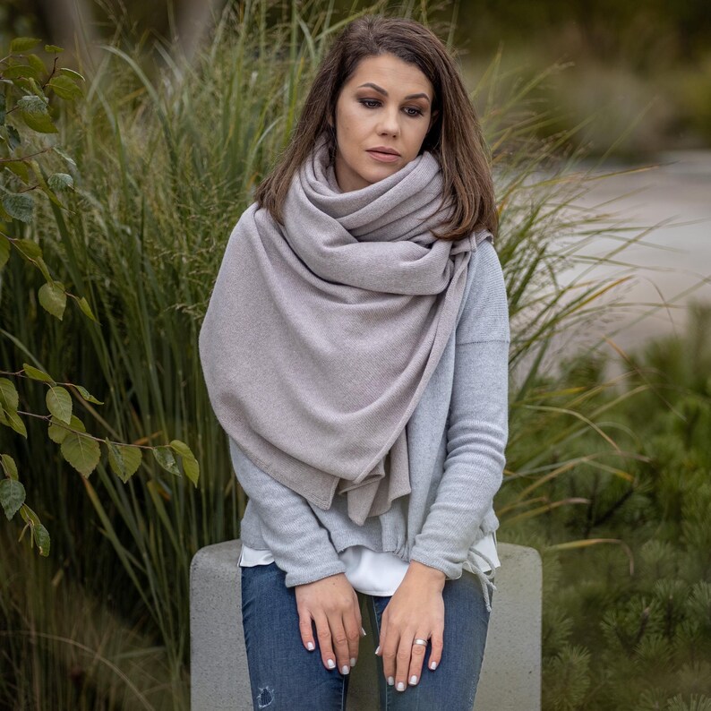 May include: A light gray scarf draped over a light gray sweater and blue jeans. The scarf is wrapped around the neck and shoulders. The background features green foliage and a concrete bench.