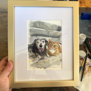 May include: Watercolor painting of a dog and cat in a gold frame. The dog is black and brown, with its tongue out. The cat is orange and white. The artwork is signed in the bottom right corner.