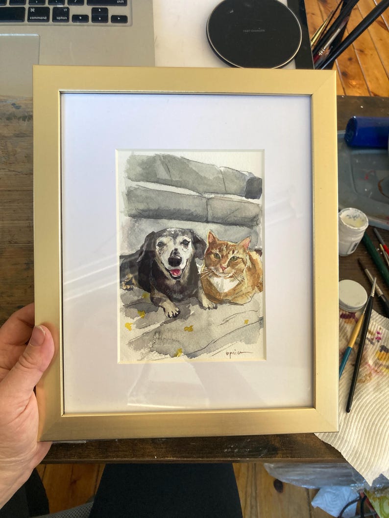 May include: Watercolor painting of a dog and cat in a gold frame. The dog is black and brown, with its tongue out. The cat is orange and white. The artwork is signed in the bottom right corner.