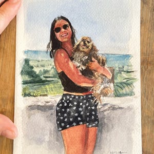 May include: Watercolor painting of a person holding a dog, with a background of a beach scene. The person is wearing sunglasses, a black top, and black shorts with white polka dots. The dog is brown and fluffy.