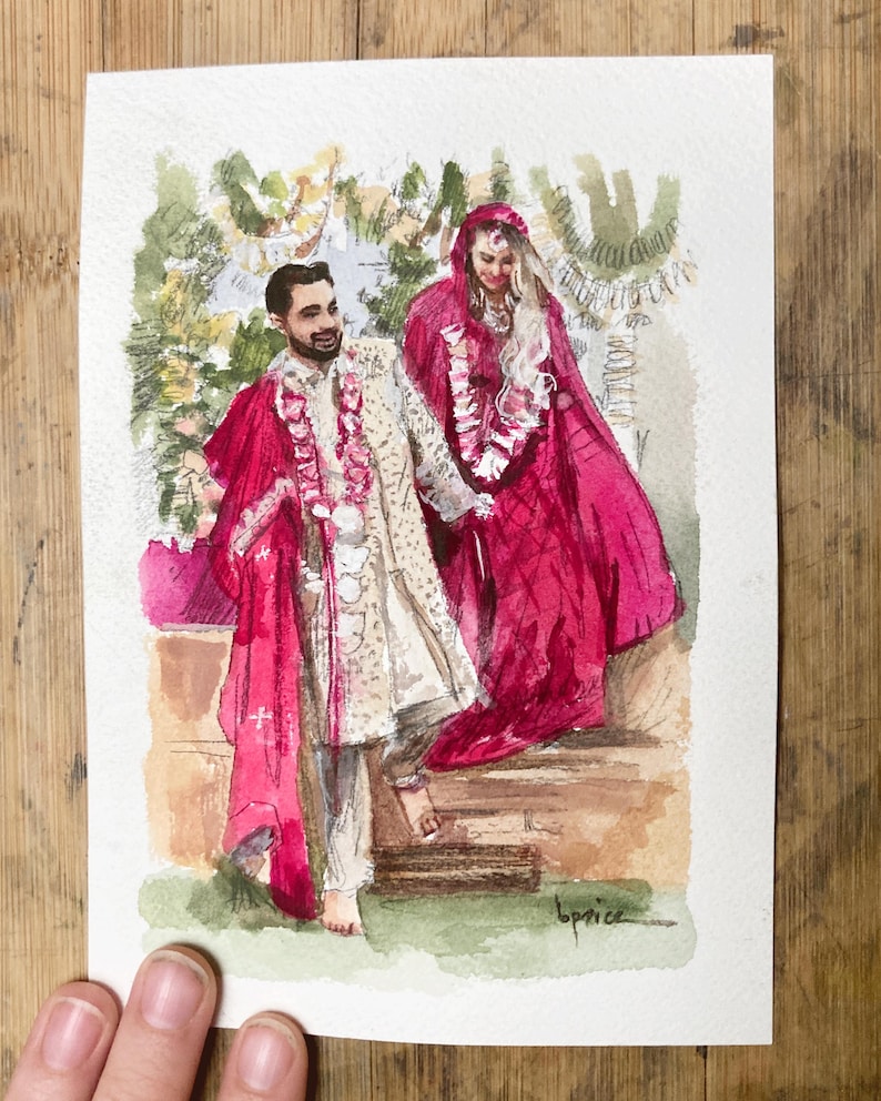 May include: Watercolor painting of a couple in traditional Indian wedding attire. The man wears a cream-colored outfit with a red shawl, and the woman is in a red sari. The artwork is on paper, with a wooden background.