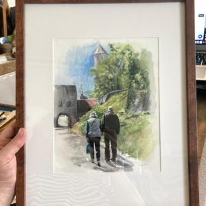 May include: Watercolor painting of a couple walking on a path towards a stone building with a tower. The artwork is framed in a brown wooden frame with a white mat. The scene includes green trees and a blue sky.
