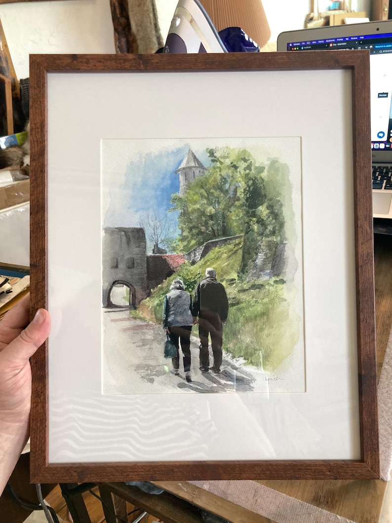 May include: Watercolor painting of a couple walking on a path towards a stone building with a tower. The artwork is framed in a brown wooden frame with a white mat. The scene includes green trees and a blue sky.