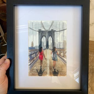 May include: Watercolor painting of two people walking on a bridge, framed in a black frame. The bridge has a distinctive arch design. The people are wearing dresses. The painting is on white matting.
