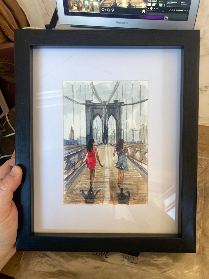 May include: Watercolor painting of two people walking on a bridge, framed in a black frame. The bridge has a distinctive arch design. The people are wearing dresses. The painting is on white matting.