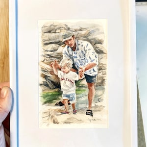 May include: Watercolor painting in a white frame. The artwork depicts a father and child walking on a rocky path. The child wears a white shirt with the word "Ballerina" and the father wears a patterned shirt. The background shows a stone wall.