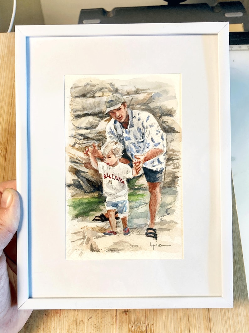 May include: Watercolor painting in a white frame. The artwork depicts a father and child walking on a rocky path. The child wears a white shirt with the word "Ballerina" and the father wears a patterned shirt. The background shows a stone wall.