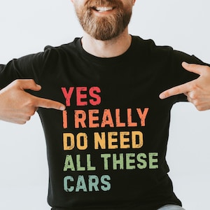 Può includere: T-shirt nera con la scritta "YES I REALLY DO NEED ALL THESE CARS" in rosso, arancione, giallo e verde. La maglietta è indossata da una persona con la barba che indica il testo.