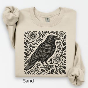 May include: A sand-coloured sweatshirt with a black and white raven illustration framed by flowers and foliage. The raven is depicted in detail. The word "Sand" is printed at the bottom of the sweatshirt.
