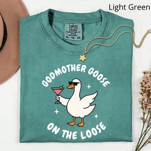 May include: Light green t-shirt with the phrase "Godmother Goose on the Loose" and a cartoon goose wearing sunglasses and holding a cocktail. A gold star pendant necklace is draped on the shirt.