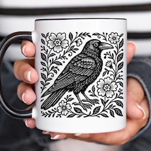 May include: White ceramic mug with a black raven illustration surrounded by floral elements. The raven is depicted in black and white, with intricate details. The mug has a black handle and rim, held by a person.