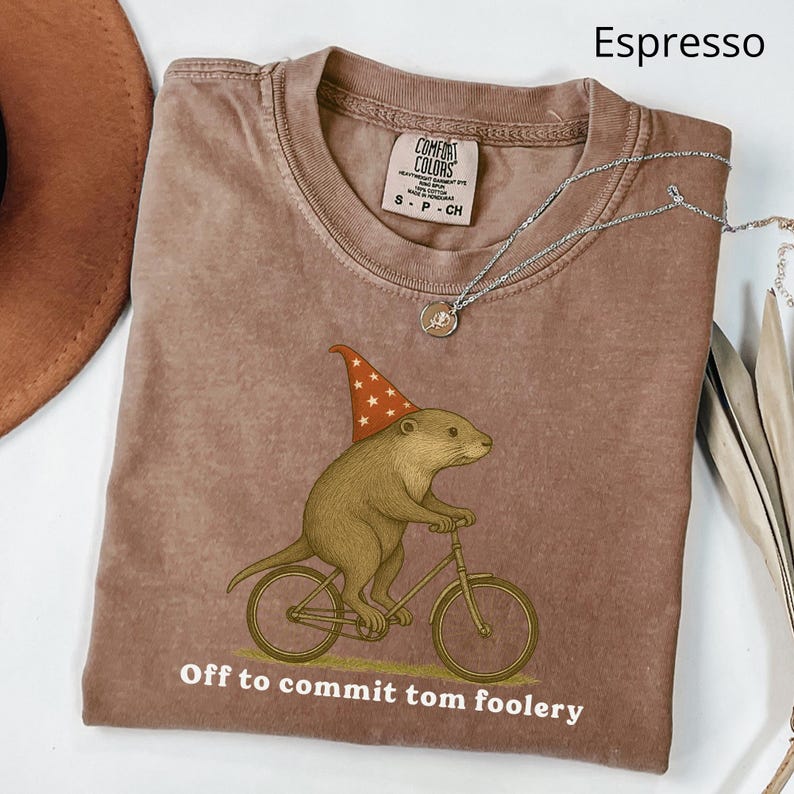 May include: Espresso-colored t-shirt with a graphic of an otter riding a bicycle and wearing a wizard hat. The text "Off to commit tom foolery" is printed below the graphic. A silver necklace is draped across the shirt.