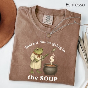 May include: Espresso-colored t-shirt with a frog chef graphic holding a spoon over a bubbling pot of soup. The text on the shirt reads "That's it. You're going in the SOUP." A silver necklace with a pendant is draped over the shirt.