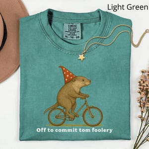 May include: Light green Comfort Colors t-shirt with a graphic of a brown rodent wearing a wizard hat and riding a bicycle. The text "Off to commit tom foolery" is below the graphic. A gold star pendant necklace is on the shirt.