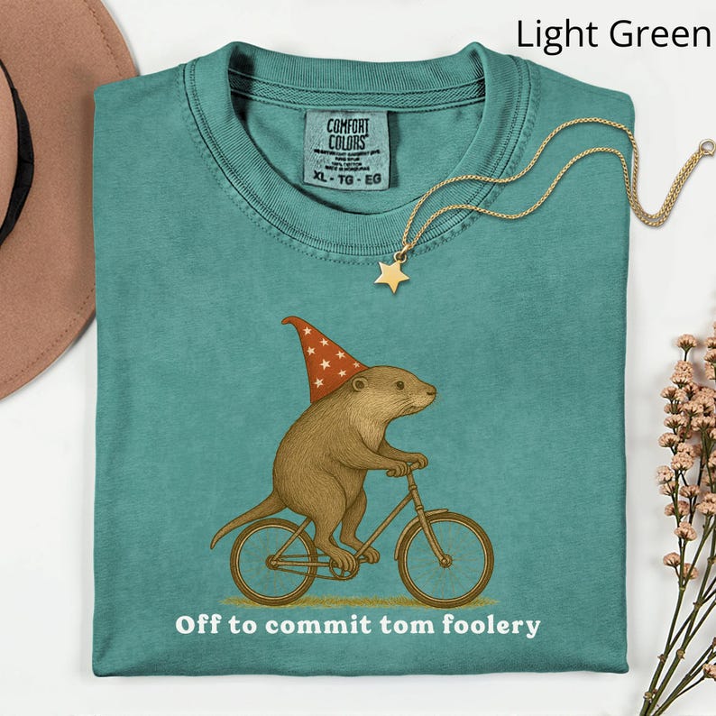 May include: Light green Comfort Colors t-shirt with a graphic of a brown rodent wearing a wizard hat and riding a bicycle. The text "Off to commit tom foolery" is below the graphic. A gold star pendant necklace is on the shirt.