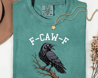 May include: Light green t-shirt with a black crow perched on a branch, the text "F-CAW-F" in white, and a gold star pendant necklace. The shirt is a Comfort Colors brand, size XL-TG-EG.