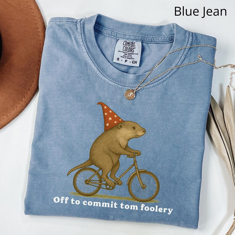 May include: A light blue t-shirt with a graphic of a brown otter wearing a wizard hat and riding a bicycle. The text "Off to commit tom foolery" is printed below the graphic. A gold necklace is draped across the shirt.