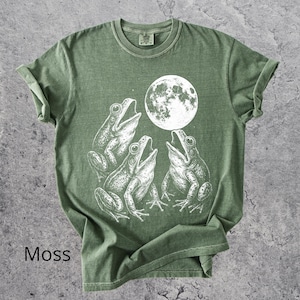 May include: Moss green t-shirt featuring a white graphic of three frogs singing under a full moon. The shirt has a classic crew neck and short sleeves. The word "Moss" is printed at the bottom left.