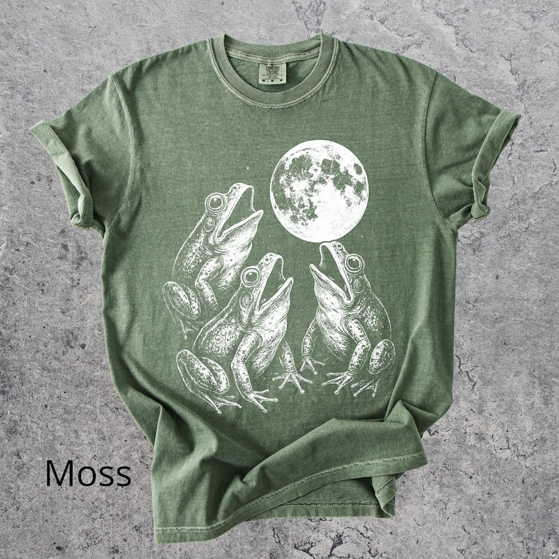 May include: Moss green t-shirt featuring a white graphic of three frogs singing under a full moon. The shirt has a classic crew neck and short sleeves. The word "Moss" is printed at the bottom left.