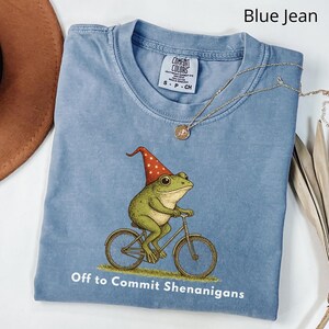 May include: Blue jean colored t-shirt with a graphic of a green frog wearing a red and white hat riding a bicycle. The text "Off to Commit Shenanigans" is printed below the graphic. A gold necklace is draped across the shirt.