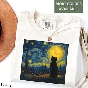 May include: Ivory-colored t-shirt featuring a black cat silhouette against a vibrant, artistic rendition of Van Gogh's "Starry Night." The shirt has a crew neck and short sleeves. The text "MORE COLORS AVAILABLE" is visible.